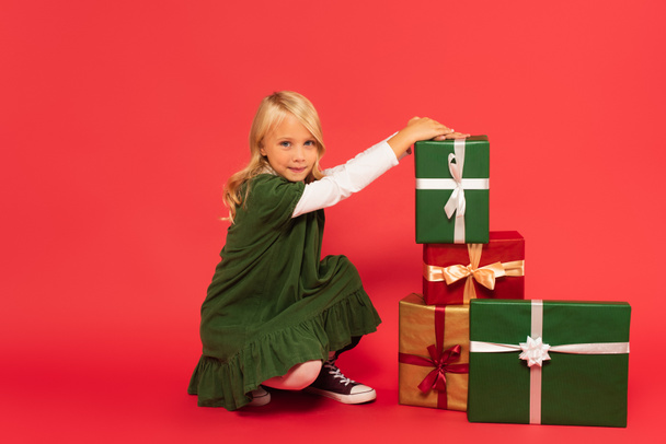 chica en vestido verde sonriendo a la cámara cerca de pila de cajas de regalo en rojo - Foto, imagen