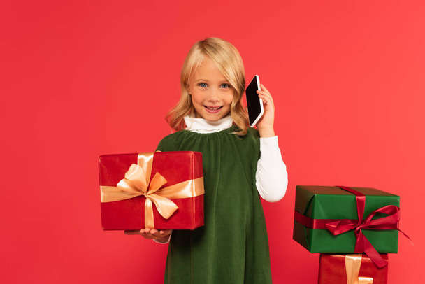 chica positiva en vestido verde hablando en el teléfono móvil cerca de regalos aislados en rojo - Foto, Imagen