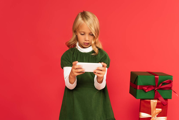 Tyttö vihreässä mekossa älypuhelimella lähellä lahjapakkauksia eristetty punaisella - Valokuva, kuva