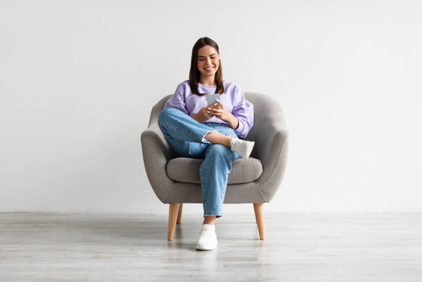 Koltukta oturan genç kadın, cep telefonu ekranına bakıyor, beyaz duvara karşı yeni bir uygulama kullanıyor. - Fotoğraf, Görsel