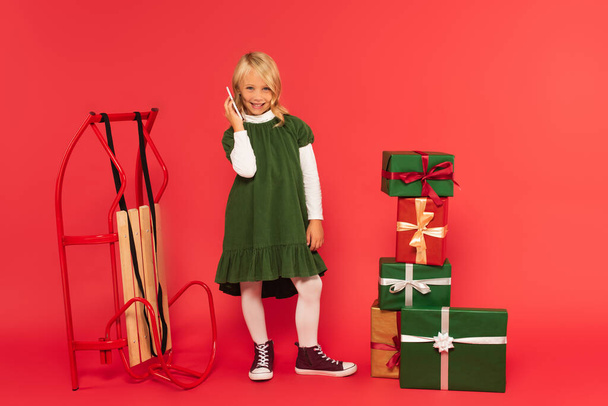smiling girl in green dress and gumshoes talking on smartphone near presents and sled on red - Photo, image