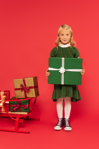 fille positive tenant boîte cadeau vert près traîneau avec des cadeaux sur rouge - Photo, image