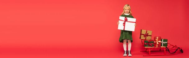 full length view of girl with big gift box near sled with presents on red, banner - Fotó, kép
