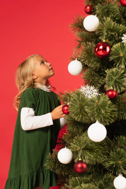girl in green dress looking at decorated christmas tree isolated on red - Foto, Imagem