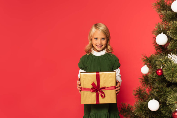 cheerful girl in green dress holding present near decorated christmas tree isolated on red - Photo, image