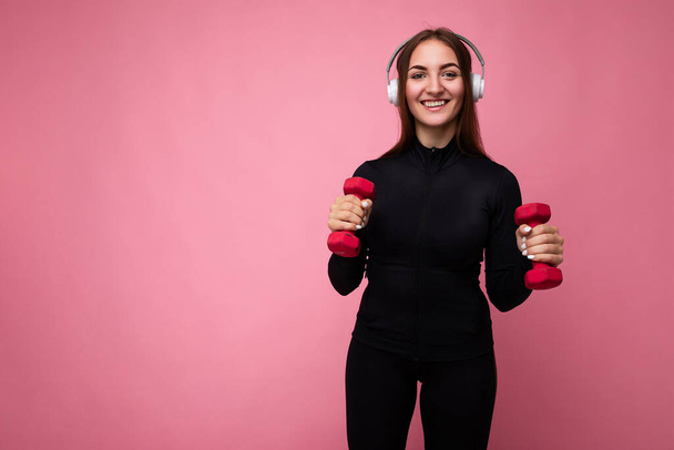 Ziemlich positiv lächelnde junge brünette Frau trägt schwarze Sportkleidung isoliert vor rosa Hintergrund Wand tun Fitness mit Hanteln tragen weiße Bluetooth-Kopfhörer hören gute Musik und - Foto, Bild