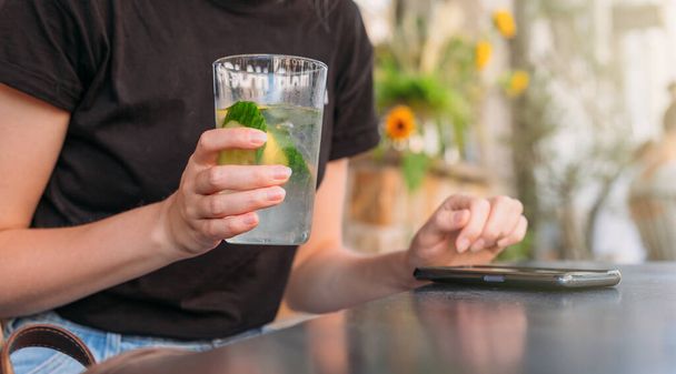 Femme au café boire de l'eau et en utilisant un téléphone mobile. Main tenant de la limonade sur fond de café. Un verre de boissons froides. Vue de la rue - Photo, image