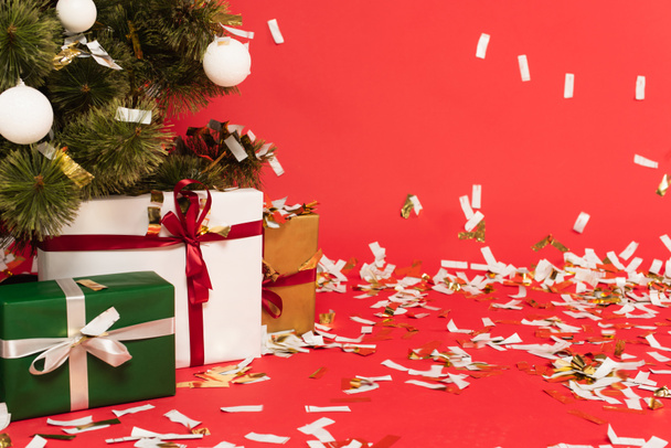 presents under decorated christmas tree near falling confetti on red - Фото, зображення