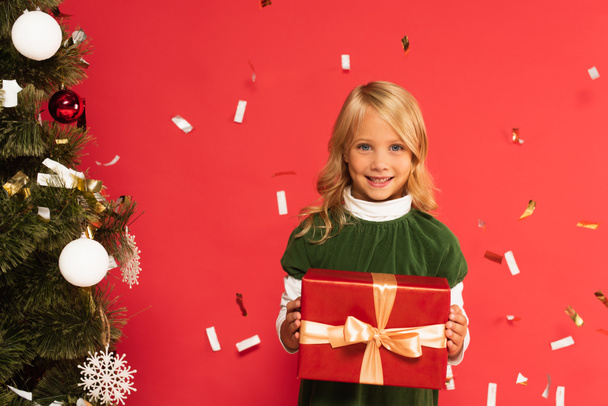 happy kid with gift box smiling at camera near christmas tree under falling confetti isolated on red - Фото, зображення