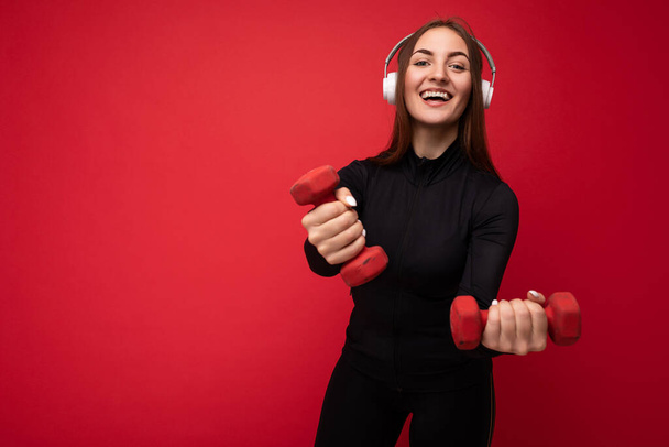 Fotografie atraktivní pozitivní úsměv mladá bruneta žena na sobě černé sportovní oblečení izolované na červeném pozadí zeď dělá fitness pomocí činky na sobě bílé bluetooth sluchátka poslouchat dobré - Fotografie, Obrázek