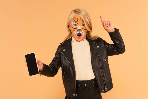 amazed girl in tiger makeup showing smartphone with blank screen and pointing with finger isolated on beige - Photo, Image