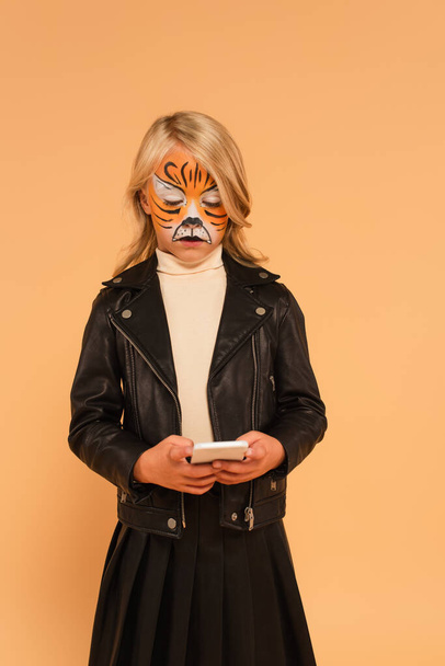 Enfant en veste en cuir noir et maquillage de tigre bavardant sur smartphone isolé sur beige - Photo, image