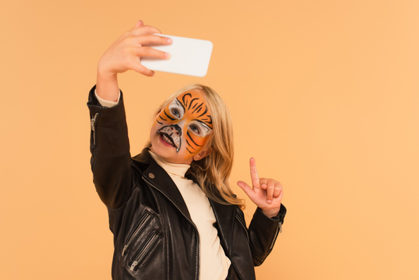 child with tiger face painting pointing up with finger while taking selfie isolated on beige - Photo, Image