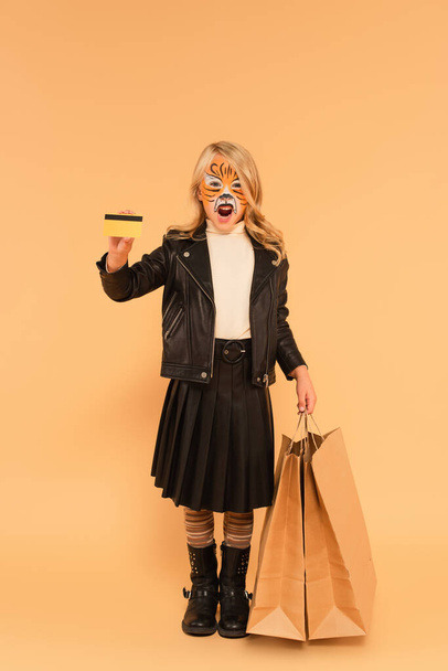 niño con pintura facial de tigre, bolsas de compras y tarjeta de crédito gruñendo en beige - Foto, Imagen