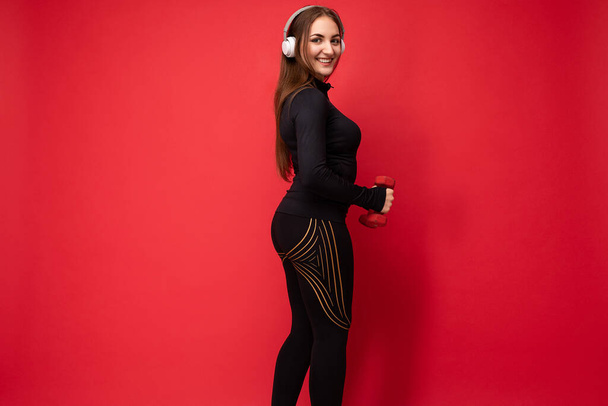 サイドプロフィールの写真魅力的な肯定的な笑顔若いブルネットの女性は白いブルートゥースイヤフォンを身に着けているダンベルを使用してフィットネスを行う赤い背景の壁に隔離された黒いスポーツ服を着て - 写真・画像
