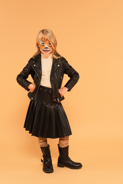 alegre chica en tigre maquillaje, chaqueta de cuero y botas posando con las manos en las caderas en beige - Foto, Imagen