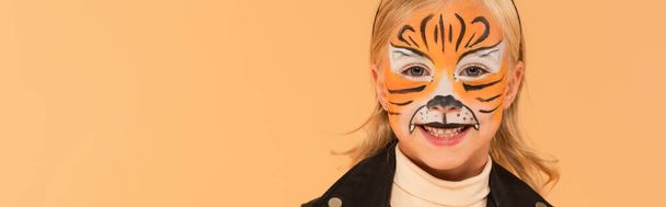 radosne dziecko z malowaniem twarzy tygrysa uśmiechnięte do kamery odizolowane na beżu, baner - Zdjęcie, obraz