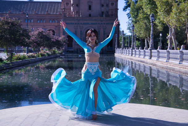 Femme hispanique d'âge moyen en robe turquoise avec strass, soulevant sa robe tout en dansant du ventre. Concept de danse du ventre. - Photo, image