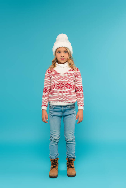 full length view of child in warm hat, jeans and sweater with ornament on blue - Foto, imagen