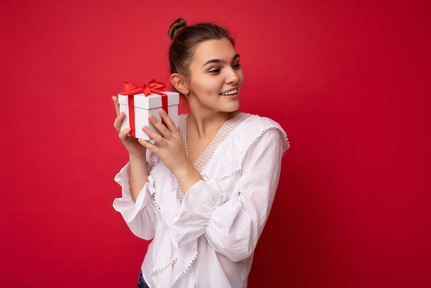 Fascynująca szczęśliwa uśmiechnięta młoda brunetka kobieta osoba odizolowana na czerwonym tle ściany noszenie białej bluzki trzymając białe pudełko z czerwoną wstążką i patrząc w bok - Zdjęcie, obraz