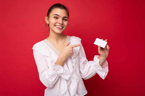 Photo of beautiful positive smiling young dark blonde woman wearing white blouse isolated over red background holding credit card looking at camera and pointing finger at plastic contactless card - Photo, Image