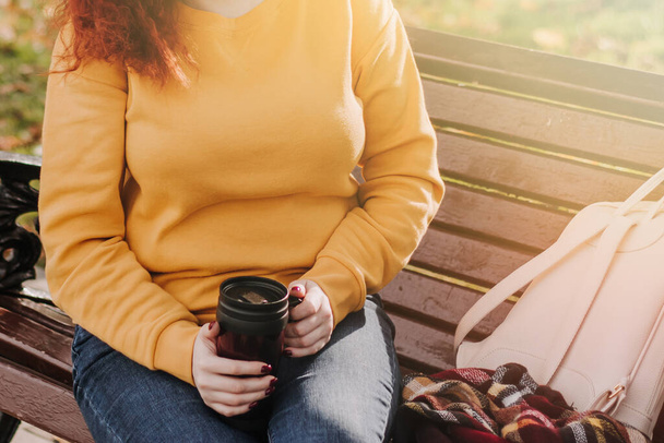 Red-haired woman in yellow sweatshirt sits on park bench and drinks coffee. She is holding reusable mug in her hands. Portrait on sunny autumn day. Lifestyle. - Photo, Image