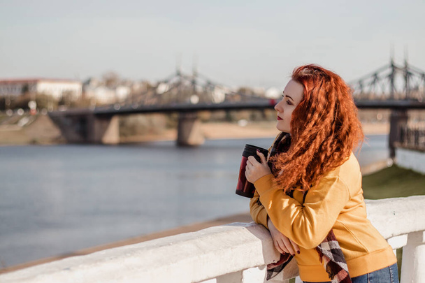 Młoda rudowłosa kobieta w żółtym swetrze trzyma kubek z kawą w rękach. Ciesz się widokiem na rzekę w słoneczny jesienny dzień. Ekologiczny styl życia, relaks. - Zdjęcie, obraz