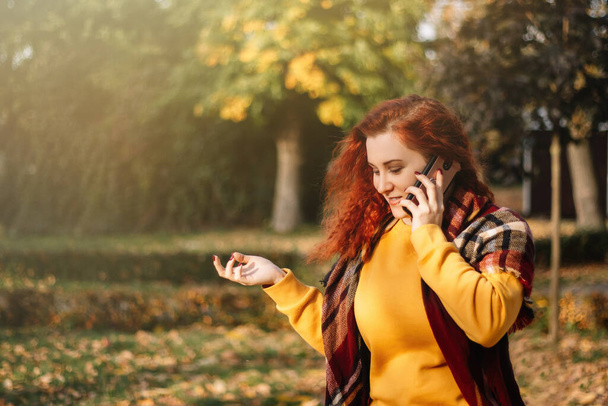 Молодая рыжая женщина в жёлтой толстовке разговаривает по телефону. Леди гуляет в осеннем парке в солнечный день. - Фото, изображение