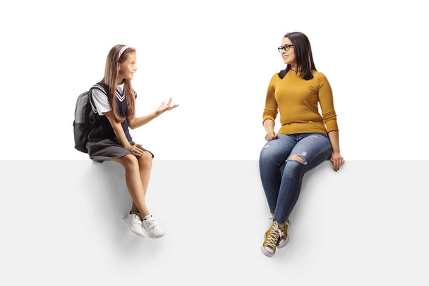 Školačka a mladá žena sedící na prázdném panelu a mající rozhovor izolované na bílém pozadí - Fotografie, Obrázek