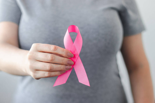 Woman in gray t-shirt holds pink ribbon in her hands. Breast Cancer Awareness Month. Supporting and promoting campaign against cancer. Health care concept. - Fotografie, Obrázek