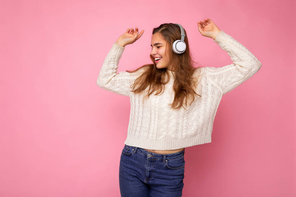 Красива щаслива усміхнена молода брюнетка кучерява жінка в білому светрі ізольована на рожевій стіні в білих навушниках Bluetooth, слухаючи холодну музику, дивлячись в бік і танцюючи
 - Фото, зображення