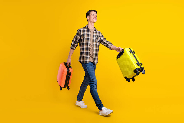 Profile photo of cheerful guy carry two bags walk wear plaid shirt jeans shoes isolated yellow color background - Photo, image