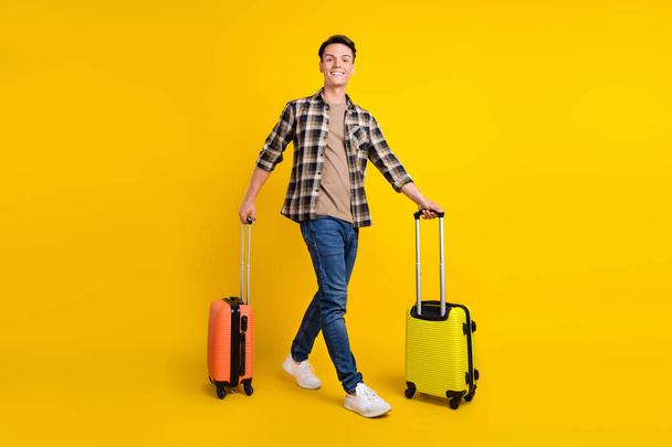 Photo of traveler guy carry two suitcase stroll wear plaid shirt jeans footwear isolated yellow color background - Zdjęcie, obraz