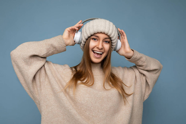 Привлекательный позитивный улыбается молодая блондинка носит бежевый зимний свитер и шляпу изолированы над синим фоном стены в белых беспроводных Bluetooth наушники слушать музыку весело и - Фото, изображение