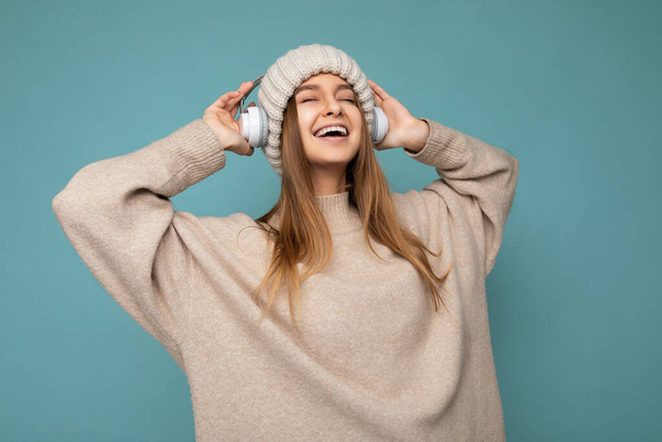 Foto di bella felice sorridente giovane donna bionda indossa maglione invernale beige e cappello isolato su sfondo blu indossando cuffie bluetooth wireless bianche ascoltare musica fresca danza e - Foto, immagini
