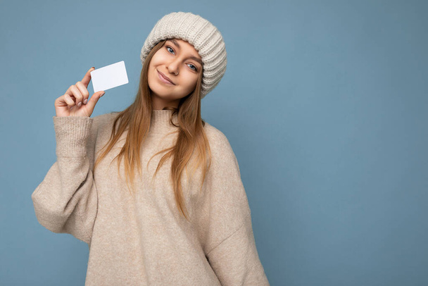 Attractive positive smiling young dark blonde woman wearing beige sweater and knitted beige hat isolated over blue background holding and showing credit card looking at camera - Photo, Image