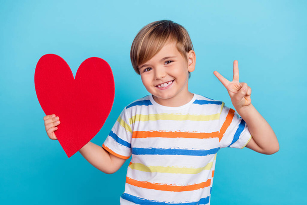 Photo of little funky blond boy hold heart wear striped t-shirt isolated on blue color background - Фото, изображение