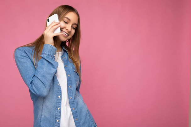 Photo of Attractive positive smiling young blonde woman wearing casual blue jean shirt isolated over pink background holding in hand and talking on mobile phone looking down - Foto, immagini