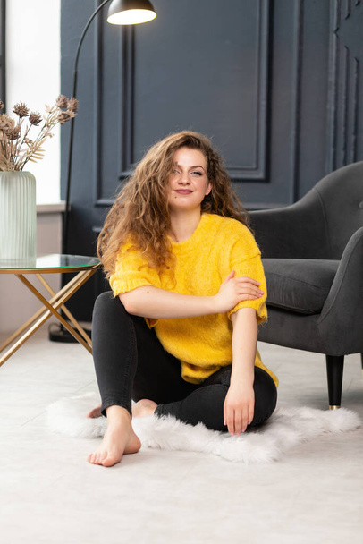 chubby girl in a stylish yellow sweater sits on the floor and smiles in a beautiful interior. Self-esteem, active life, charming plus-size, positive body. - Zdjęcie, obraz
