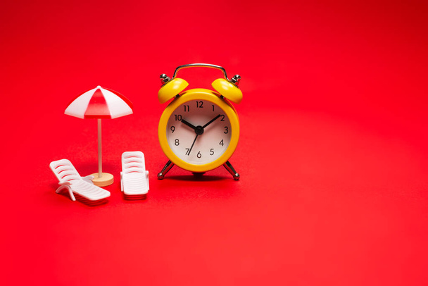 Time to travel. Sun lounger, umbrella and alarm clock on a red background. - Photo, Image