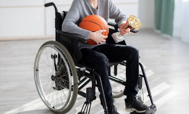 Jogador de basquete adolescente irreconhecível com bola e troféu sentado em cadeira de rodas, sofrendo por lesão em casa - Foto, Imagem