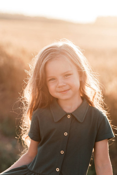 Belle petite fille aux cheveux longs marchant à travers un champ de blé par une journée ensoleillée. Portrait extérieur. Enfants relaxants - Photo, image