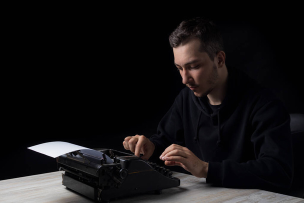 Młody przystojny mężczyzna w czarnym kapturze pisze na maszynie do pisania, na czarnym tle w niskim tonem - Zdjęcie, obraz