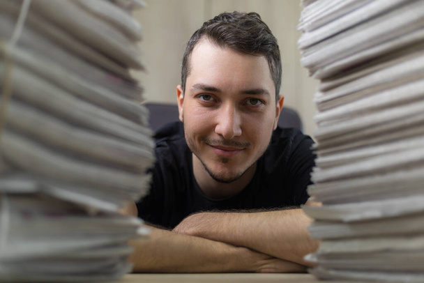 Close up portrait of a young journalist and a stacks of newspapers in a blurred foreground. - Photo, Image
