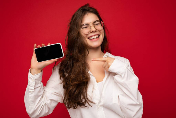 Улыбающийся счастливый привлекательный молодой брюнет женщина в белой рубашке и оптические очки изолированы на красном фоне, держа в руке и показывая мобильный телефон с пустым экраном для макета - Фото, изображение