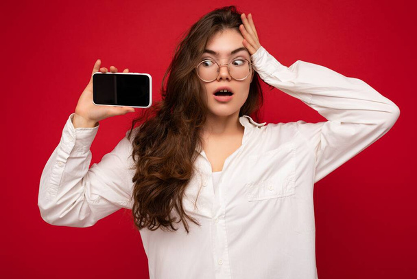 fotografie sexy ohromen krásná mladá bruneta žena na sobě bílou košili a optické brýle izolované přes červené pozadí drží v ruce a ukazuje mobilní telefon s prázdnou obrazovkou pro výstřižek hledá - Fotografie, Obrázek