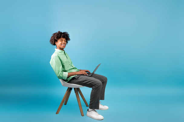 Koncepcja e-learningu. czarny facet student za pomocą laptop komputer siedząc w krzesło nad niebieski studio tło, puste miejsce - Zdjęcie, obraz