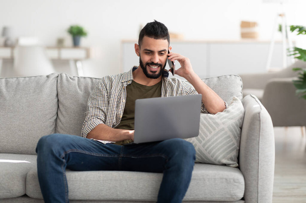 Onnellinen nuori arabi mies käyttää kannettavaa tietokonetta videoneuvotteluun, soittamalla älypuhelimeen, työskentelemällä verkossa kotoa - Valokuva, kuva