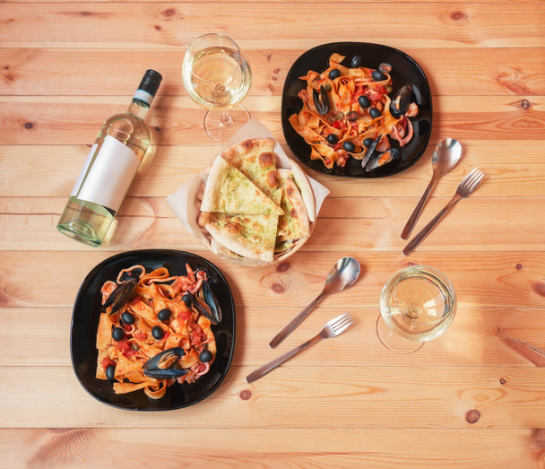 Platen pappardelle pasta met zeevruchten met tomatensaus, fles witte wijn, glazen witte wijn en focaccia brood op houten tafel. Bovenaanzicht. - Foto, afbeelding