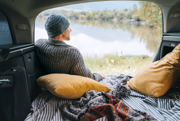 A man dressed in warm knitted clothes sitting in the cozy camper trunk and enjoying the mountain lake view. Warm early autumn auto traveling concept inside car pilows and blankets view image.   - Valokuva, kuva